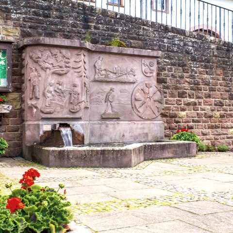 Ein steinerner Dorfbrunnen mit Reliefs und fließendem Wasser, umgeben von roten Blumen und einer Infotafel an einer Steinmauer., © TI Bitburger Land - M. Mayer