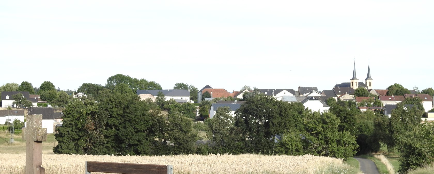 Een bank en een kruis staan voor een veld. Op de achtergrond is een dorp te zien met een kerk en twee torens., © Thomas Neises