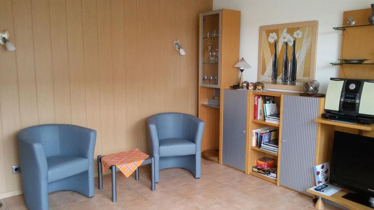 Ein gemütlicher Wohnraum mit zwei blauen Sesseln und einem kleinen Tisch. Im Hintergrund befinden sich Bücherregale und dekorative Elemente.