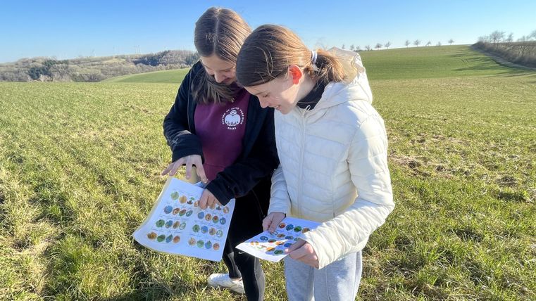 Zwei Mädchen stehen auf einem Feld und betrachten aufmerksam einige Blätter mit Abbildungen. Im Hintergrund erstreckt sich eine grüne Landschaft unter einem klaren Himmel.