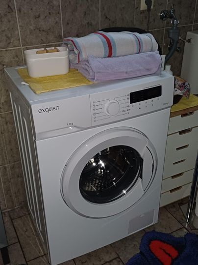 A washing machine stands in a bathroom. Towels are neatly placed on it and the walls are tiled.