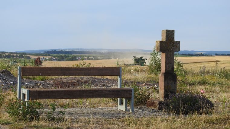 Bank und Steinkreuz auf einem Feldweg mit Mähdrescher im Hintergrund.