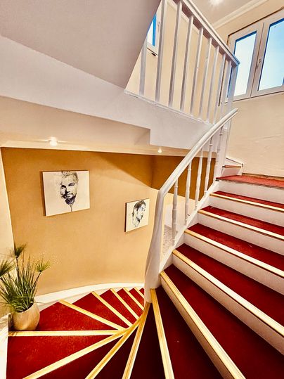 A bright staircase with red steps and a white railing. Two portraits hang on the wall, and a plant stands at the bottom.