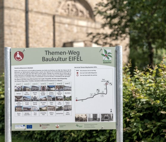 Panneau du sentier thématique Baukultur Eifel à Wolsfeld avec carte et informations sur les curiosités. On peut voir un bâtiment à l'arrière-plan., © Naturpark Südeifel, Thomas Urbany