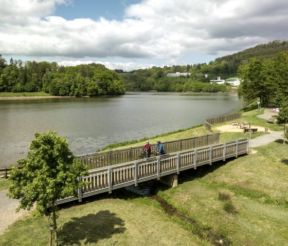 Zwei Radfahrer überqueren eine Holzbrücke am Stausee Bitburg, umgeben von grüner Landschaft und bewölktem Himmel., © Eifel Tourismus GmbH, Dominik Ketz