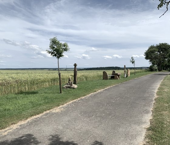 Asphaltierter Weg durch Felder, mit Bänken und Steinskulpturen am Rand, unter blauem Himmel mit Wolken., © B. Milbach