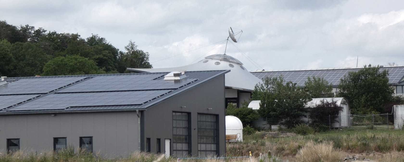 Ein modernes Gebäude mit Solarpanelen auf dem Dach, im Hintergrund eine UFO-ähnliche Struktur mit Antenne, umgeben von Bäumen und Wiese., © TI Bitburger Land