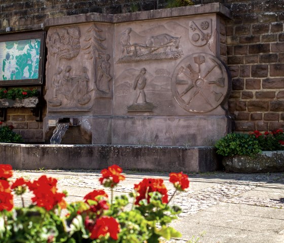 Ein Dorfbrunnen mit kunstvollen Reliefs aus Stein, umgeben von roten Blumen. Eine Infotafel und eine Bank sind ebenfalls sichtbar., © TI Bitburger Land M. Mayer