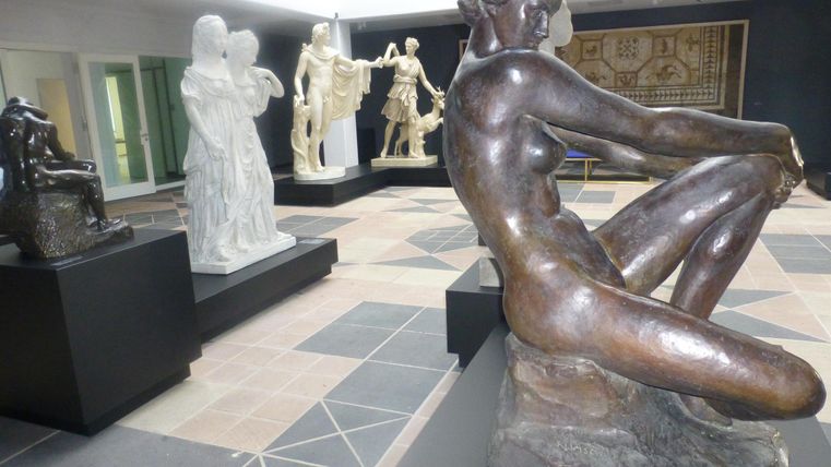 In einem modernen Ausstellungsraum sind mehrere Statuen aus verschiedenen Materialien zu sehen. Im Vordergrund steht eine bronzene Figur einer sitzenden Frau, umgeben von weiteren Skulpturen.