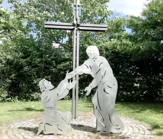 Sculpture en métal avec une croix et deux personnages, l'un agenouillé, l'autre debout, sur un cercle pavé dans un jardin verdoyant.