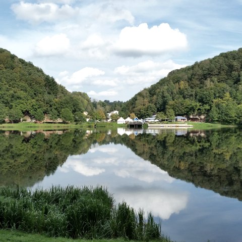 Panorama of the Bitburg reservoir with wooded hills, buildings on the shore and a clear reflection in the water under a blue sky., © TI Bitburger Land