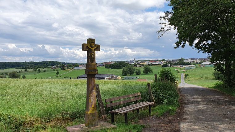 Wegekreuz und Bank mit Blick auf Mötsch, umgeben von grünen Feldern und einem bewölkten Himmel.