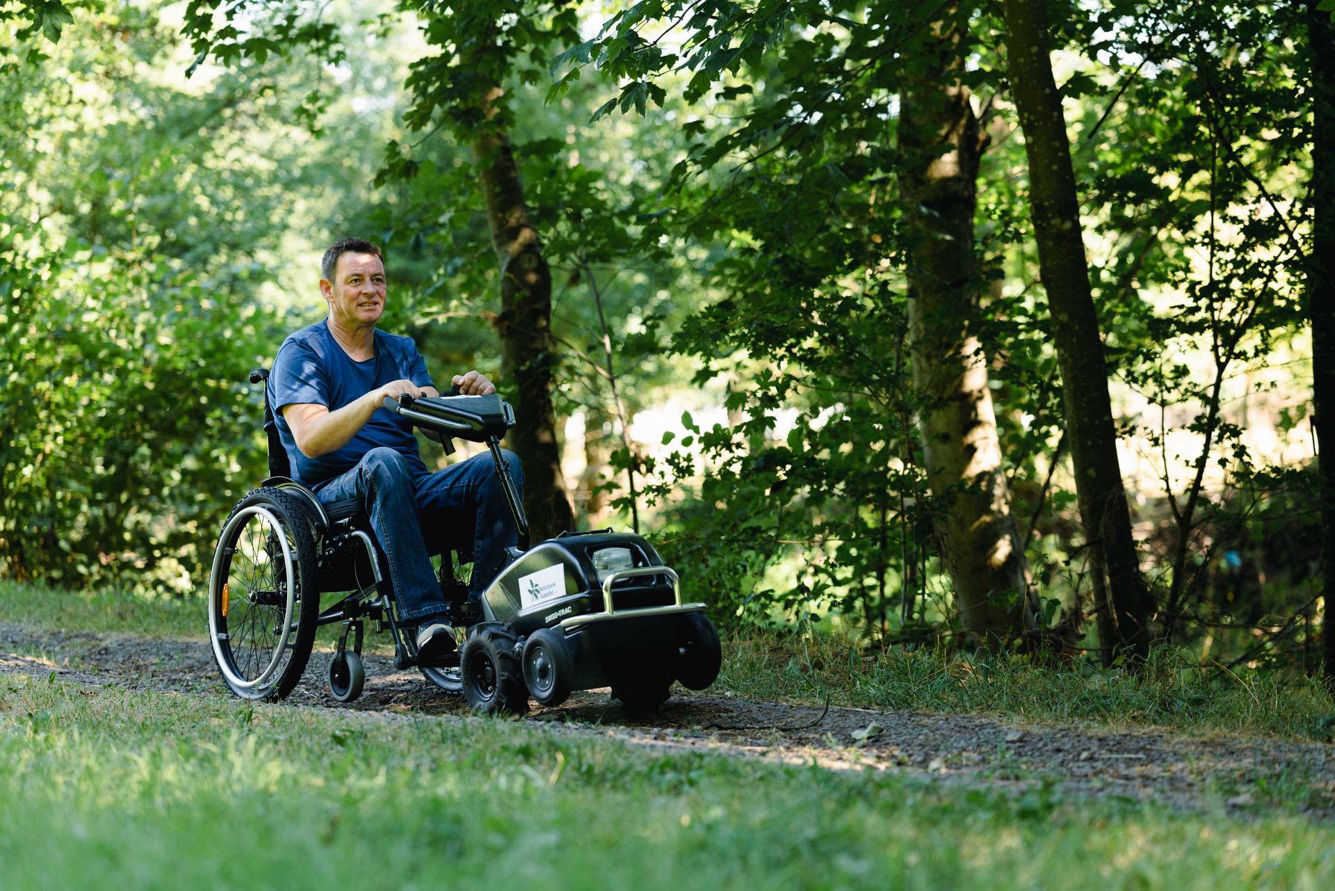 Ein Mann im Rollstuhl mit Rollstuhlzuggerät fährt über einen Waldweg