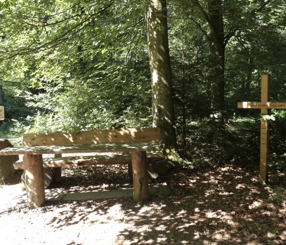 Eine Holzbank steht im Wald neben einem Kreuz. Ein Wegweiser zeigt in Richtung Speicher. Sonnenlicht fällt durch die Blätter., © TI Bitburger Land