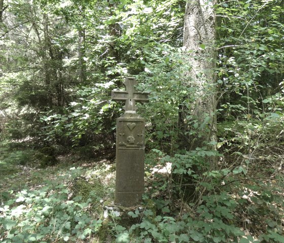 Ein Steinkreuz steht im Wald, umgeben von dichtem Laub und Bäumen. Der Boden ist mit Moos und Blättern bedeckt., © TI Bitburger Land
