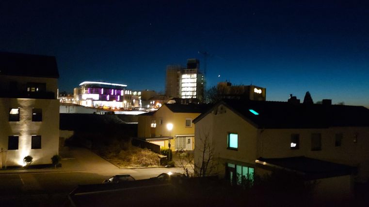 Eine städtische Szene bei Nacht mit beleuchteten Häusern und einem klaren Himmel. Im Hintergrund sind moderne Gebäude und ein sanfter Farbverlauf sichtbar.