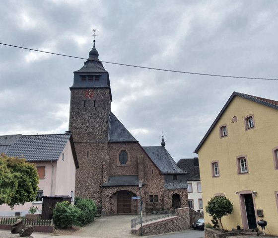 Eine Kirche in Gransdorf mit einem markanten Turm, umgeben von Wohnhäusern und Bäumen unter einem bewölkten Himmel., © TI Bitburger Land S.Wagner
