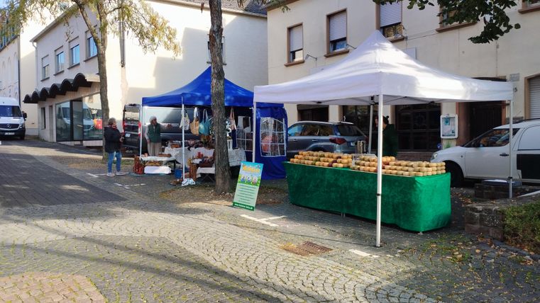 Un stand de marché avec des tentes bleues et blanches se trouve le long d'une route. Des gens sont visibles tandis qu'ils achètent des produits frais et des aliments.