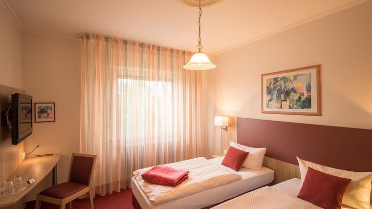 Ein gemütliches Zimmer mit zwei Einzelbetten und einem Schreibtisch. Große Fenster mit Vorhängen lassen viel Licht herein.