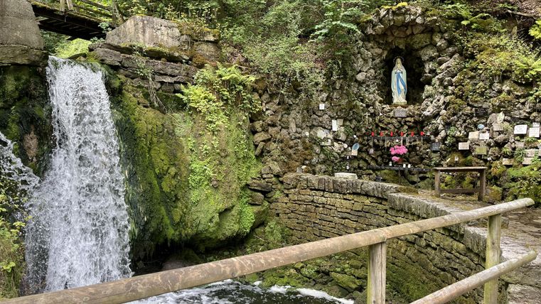 Ein schöner Wasserfall fließt neben einer steinernen Wand mit einer Statue von Maria. Umgeben von üppigem Grün und Natur, lädt der Ort zur Ruhe ein.