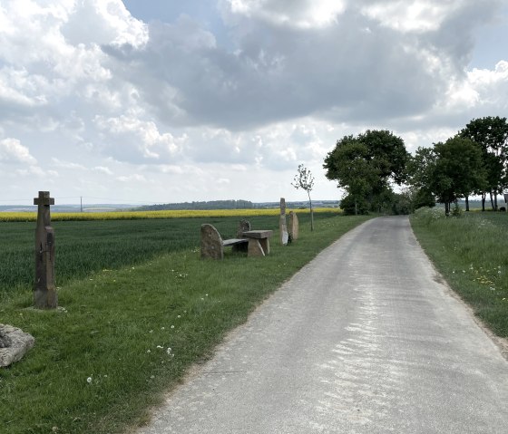 Un chemin rural passe devant une croix en pierre et un banc, entourés de champs et d'arbres verts sous un ciel nuageux., © B. MIlbach