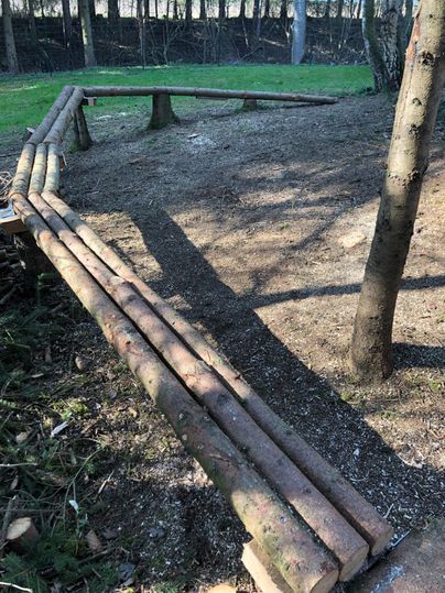 A curved wooden path made of tree trunks in a forest. The ground is covered with soil and small plants.