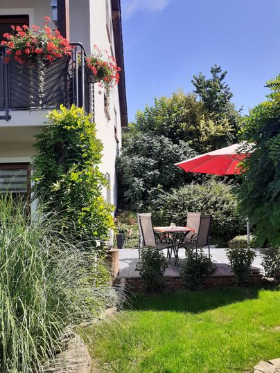 Een gezellige tuin met een terras. Een tafel met stoelen staat onder een rode parasol, omringd door groene planten.