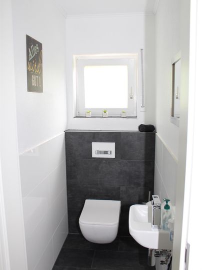 Une salle de bain moderne avec des toilettes blanches et un petit lavabo. Les murs sont partiellement carrelés en clair et en foncé, et il y a une fenêtre qui laisse entrer la lumière naturelle.