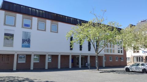 Een modern gebouw met grote ramen en een entree. Op de voorgrond staat een boom, en de lucht is helder en blauw.