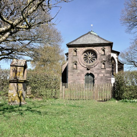 De Frohnertkapel in Oberkail, omringd door bomen en een oude grafsteen op de voorgrond, bij zonnig weer., © TI Bitburger Land