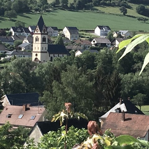Dorfansicht mit Kirche im Zentrum, umgeben von Häusern und grünen Hügeln. Im Vordergrund sind Pflanzen zu sehen., © TI Bitburger Land