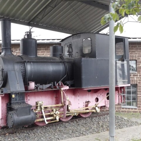 Een oude stoomlocomotief staat onder een afdak voor een bakstenen gebouw. De locomotief is zwart en roze geschilderd., &copy; Eifelverein Ortsgruppe Speicher