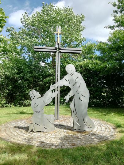 Metallskulptur mit Kreuz und zwei Figuren in einem Garten.