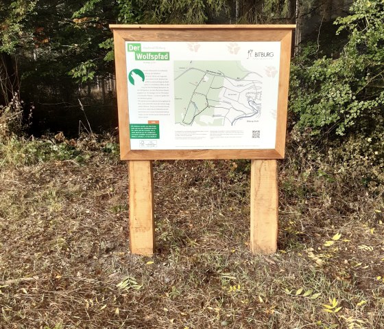 Ein Holzschild im Wald zeigt eine Karte des Wolfspfads in Bitburg. Es steht auf einer Wiese, umgeben von Bäumen und Laub., © Otmar Koch