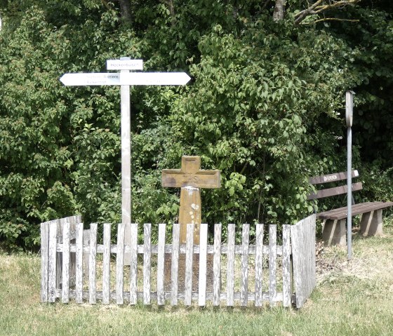 Un panneau indicateur et une croix en bois se trouvent sur un sentier de randonnée, entourés de buissons verts. Un banc se trouve à côté., © TI Bitburger Land