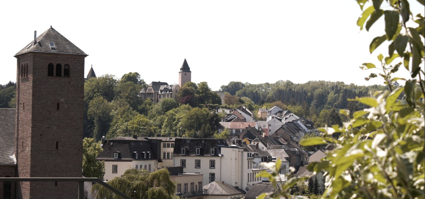 Panoramic view of Kyllburg with a church tower in the foreground, surrounded by houses and green trees under a blue sky., © TI Bitburger Land
