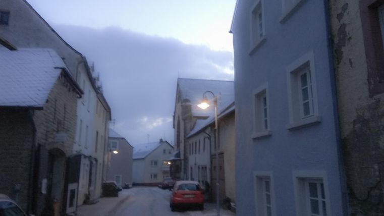 Eine ruhige Straße in einem kleinen Dorf, umgeben von Schnee und alten Gebäuden. Die Dämmerung sorgt für eine gemütliche Atmosphäre.