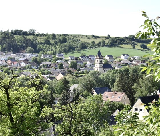 Vue panoramique du village d'Oberweis avec une église au centre, entouré d'un paysage verdoyant et d'arbres au premier plan., © TI Bitburger Land