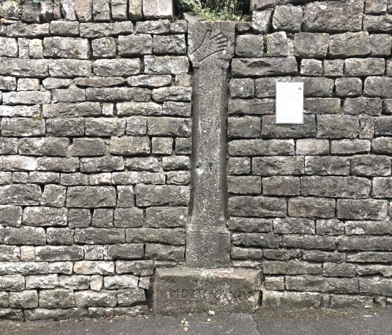 Eine alte Steinmauer mit einer Kreuzwegstation und einer Infotafel. Die Station zeigt ein Relief einer Hand. Darunter ist eine Inschrift., © TI Bitburger Land