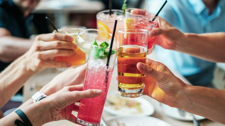 Several hands hold different cocktails as they toast over the table. In the background, food and guests can be seen, creating a cozy atmosphere.