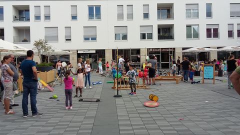 Un terrain de jeu animé avec des enfants participant à diverses activités. Entouré de bâtiments résidentiels modernes et de personnes qui regardent ou participent.