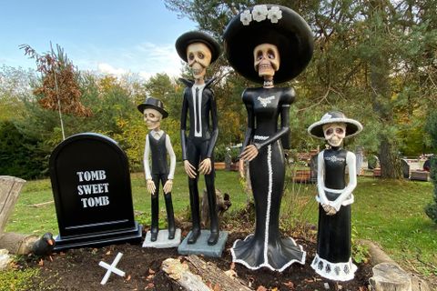 A group of four skeleton figures in festive clothing stands around a tombstone. The tombstone bears the inscription "TOMB SWEET TOMB" and the surroundings are adorned with plants.