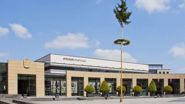 Een modern gebouw met grote ramen en een lichte gevel. Op de voorgrond bevinden zich beplante gebieden en een groene sculptuur.