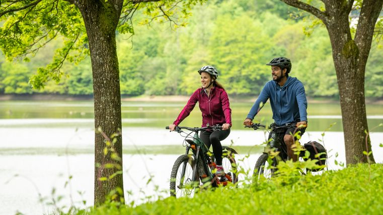 Zwei Personen fahren auf Fahrrädern entlang eines Sees, umgeben von grüner Natur.