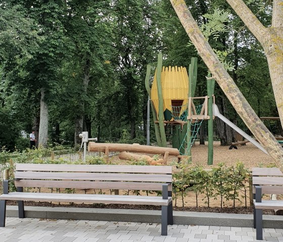 Speeltuin in het Maximiner Wäldchen park in Bitburg met bankjes, klimrek en glijbaan, omgeven door bomen en groen., © TI Bitburger Land