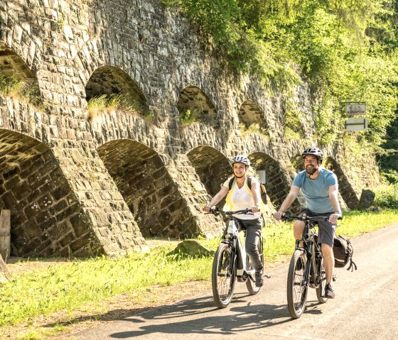 Zwei Radfahrer fahren auf einem sonnigen Weg entlang einer alten Steinmauer mit Bögen, umgeben von grüner Vegetation., © Eifel Tourismus GmbH, Dominik Ketz