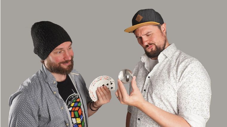 Twee mannen staan voor een grijze achtergrond. De man links houdt een rond object en een wit iets vast, terwijl de man rechts sceptisch naar een zilveren voorwerp kijkt.