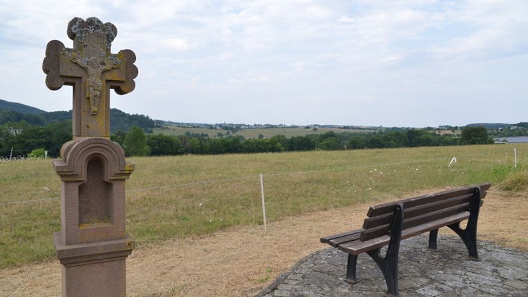 Eine Bank und ein steinernes Kreuz in einer ländlichen Landschaft mit weitem Blick über Felder und Hügel.