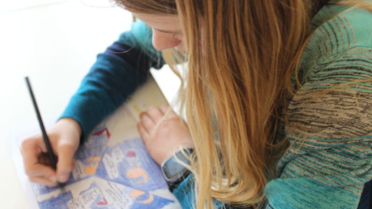 Ein Mädchen mit langen Haaren zeichnet konzentriert auf einem Blatt Papier. Sie trägt einen grünen Pullover und hält einen Bleistift in der Hand.