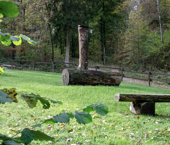 Wooden sculpture and bench on a green meadow, surrounded by forest. A hiking trail leads through the autumnal landscape., © Tourist-Info Bitburger Land